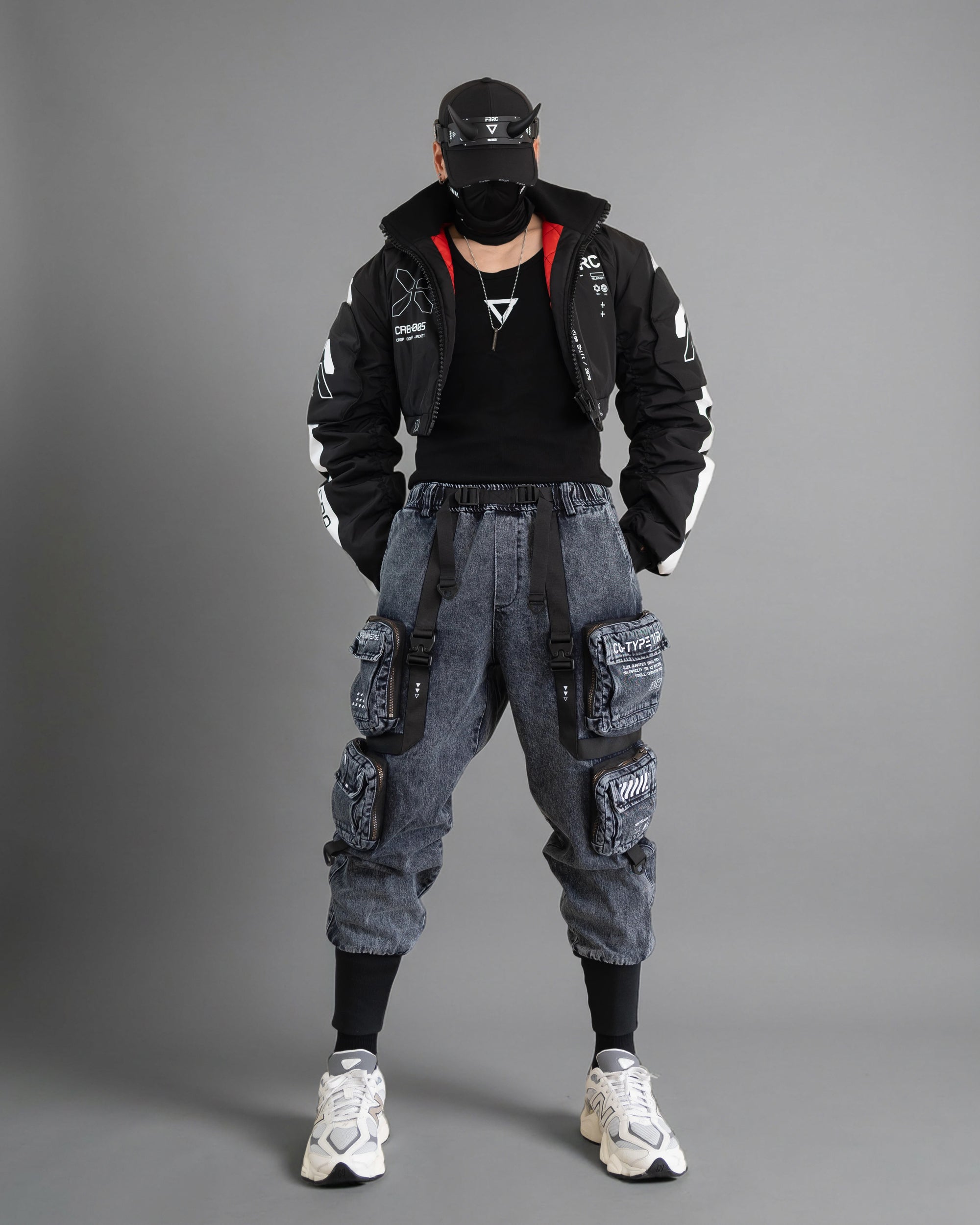 Full cyberpunk outfit with a cropped black bomber, technical markings, strapped cargo trousers, face covering and futuristic sneakers.